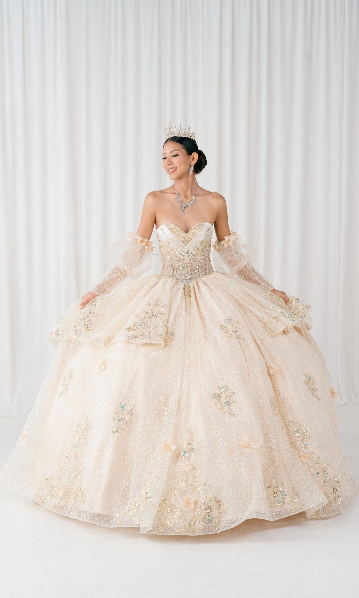 A woman in the Champagne Quinceanera Dress 8385J by Cinderella Couture, with a jeweled tiara and necklace, poses in front of white curtains. The dress features a corset bodice, floral embellishments, and a full skirt.